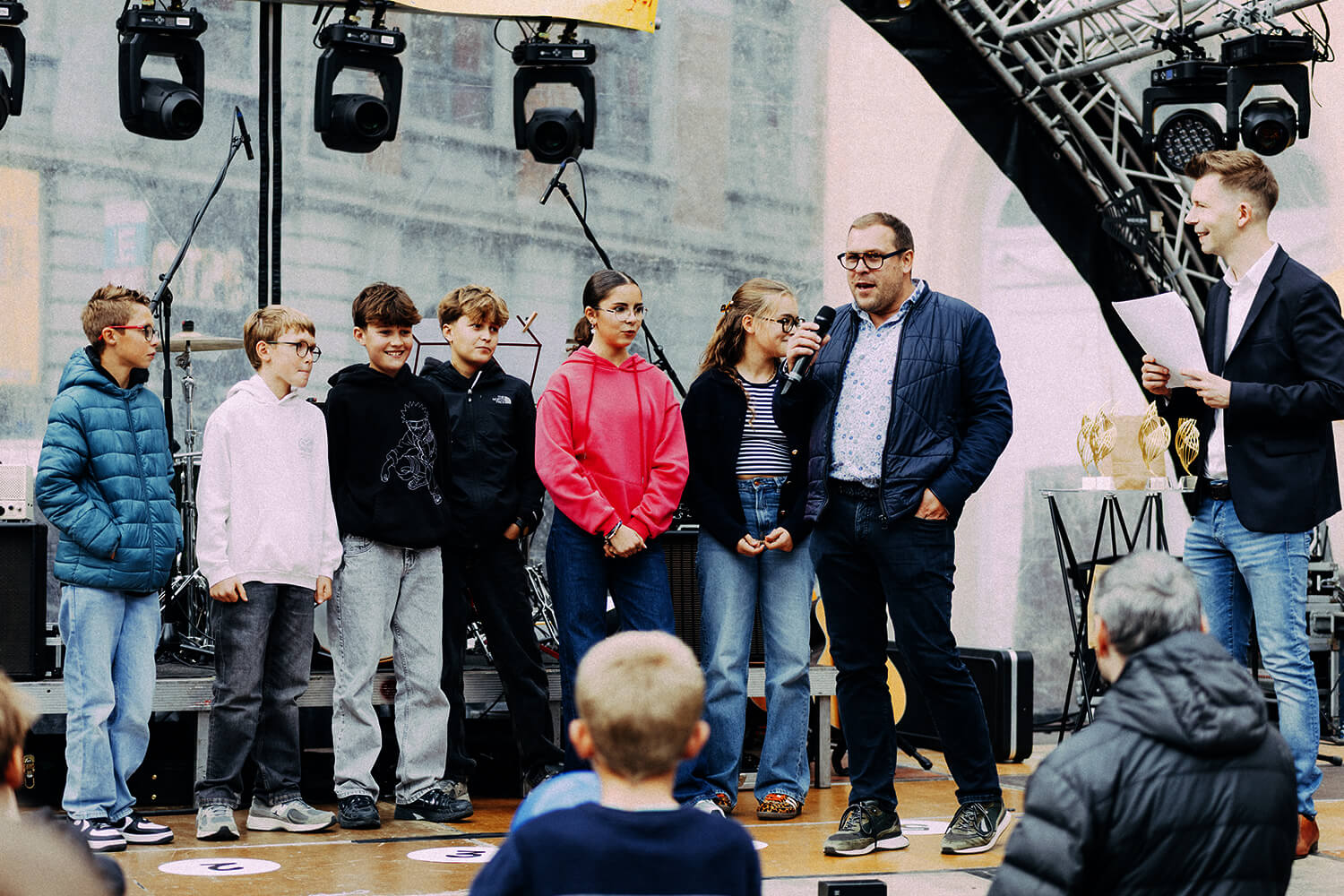 Six jeunes et leur coach sur scène, lors d'une présentation publique. L'un des adultes parle au micro, tandis que l'autre tient une feuille. Le public, dont des enfants, écoute attentivement.