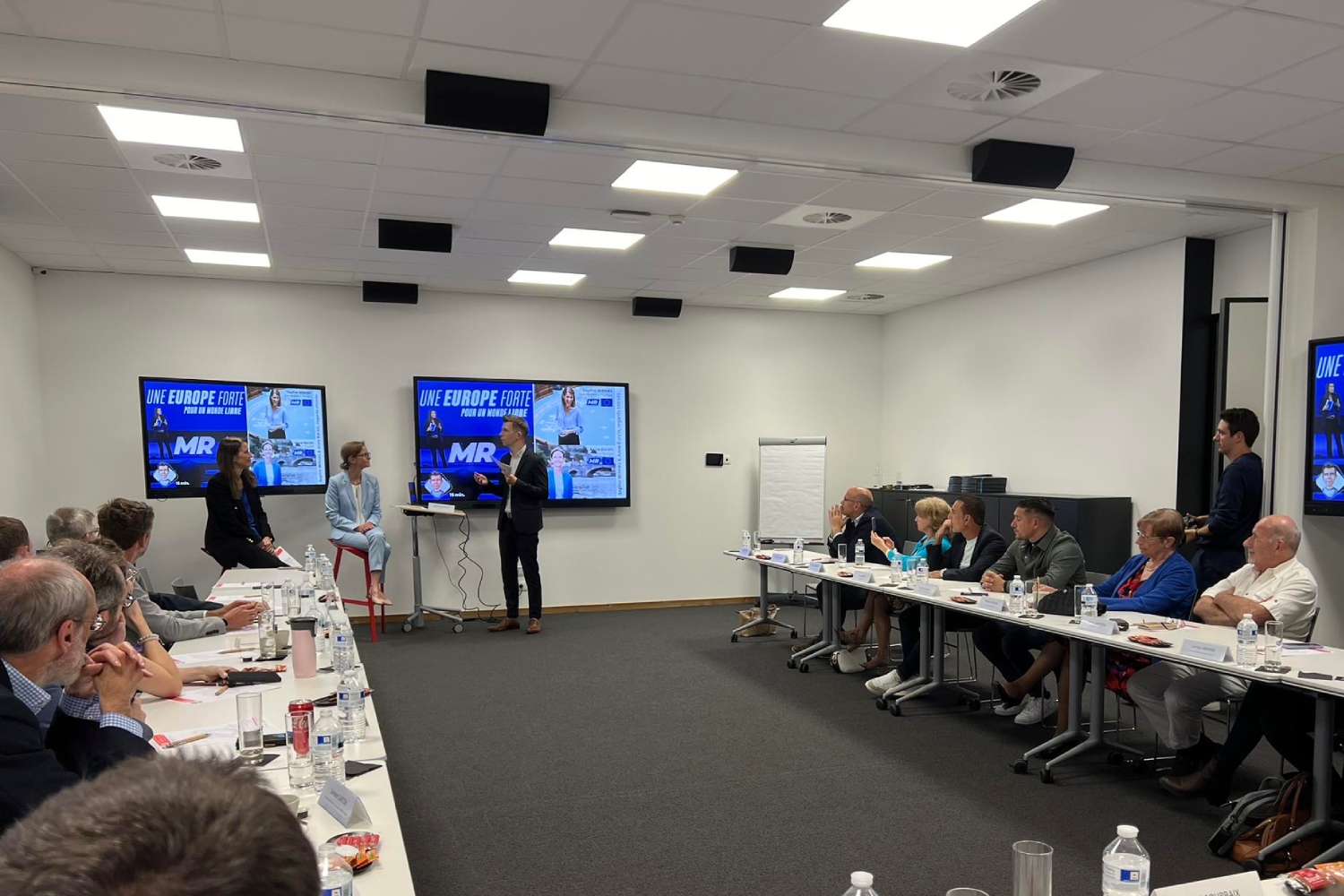 Nicolas Pourbaix interrogeant Sophie Wilmès et Anne Barzin face aux participant-e-s présent-e-s qui semblent à l'écoute.