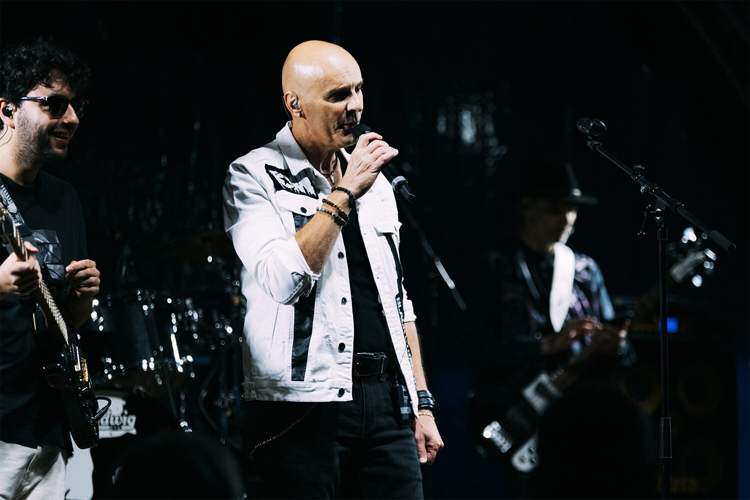 Le chanteur "FAN – Tribute Pascal Obispo" en veste blanche, au centre de la scène, interprète avec passion un morceau au micro. À sa gauche, un guitariste sourit en jouant, tandis qu’à droite, un claviériste se concentre sur son instrument.