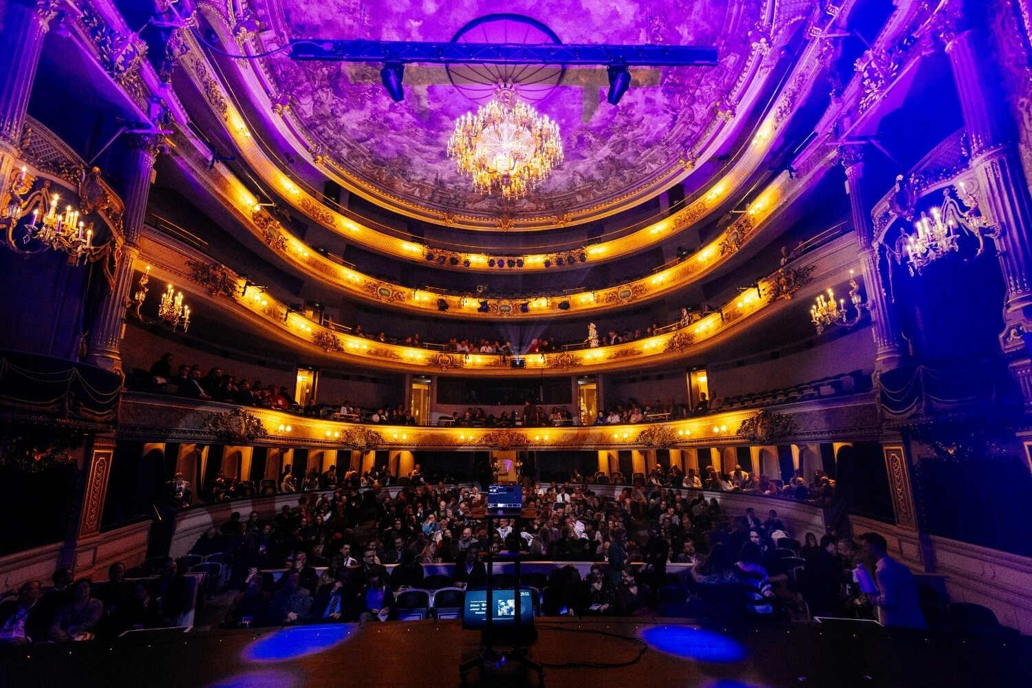 Photo d&rsquo;ambiance de la salle de spectacle : vue d&rsquo;ensemble du Th&eacute;&acirc;tre royal de Namur, balcons &eacute;clair&eacute;s de lumi&egrave;res dor&eacute;es et violettes, public install&eacute; avant une conf&eacute;rence ou un spectacle.