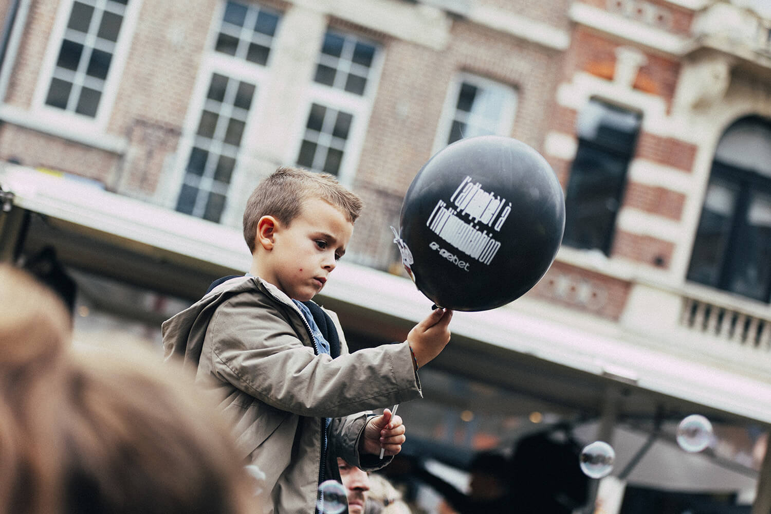 Un enfant tient un ballon noir avec un logo blanc lors d'un événement en extérieur. Il regarde attentivement le ballon qu'il tient devant lui. En arrière-plan, des bâtiments et des passants sont visibles.
