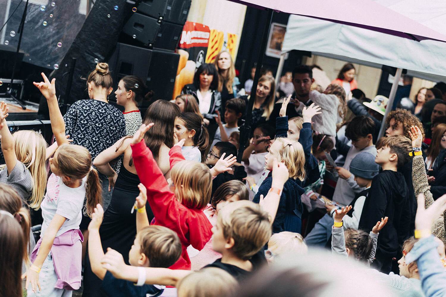 Une foule d'enfants, les mains levées, applaudit ou participe à une activité devant une scène extérieure. Des adultes sont également présents et semblent encourager les enfants. L'ambiance est festive et dynamique.