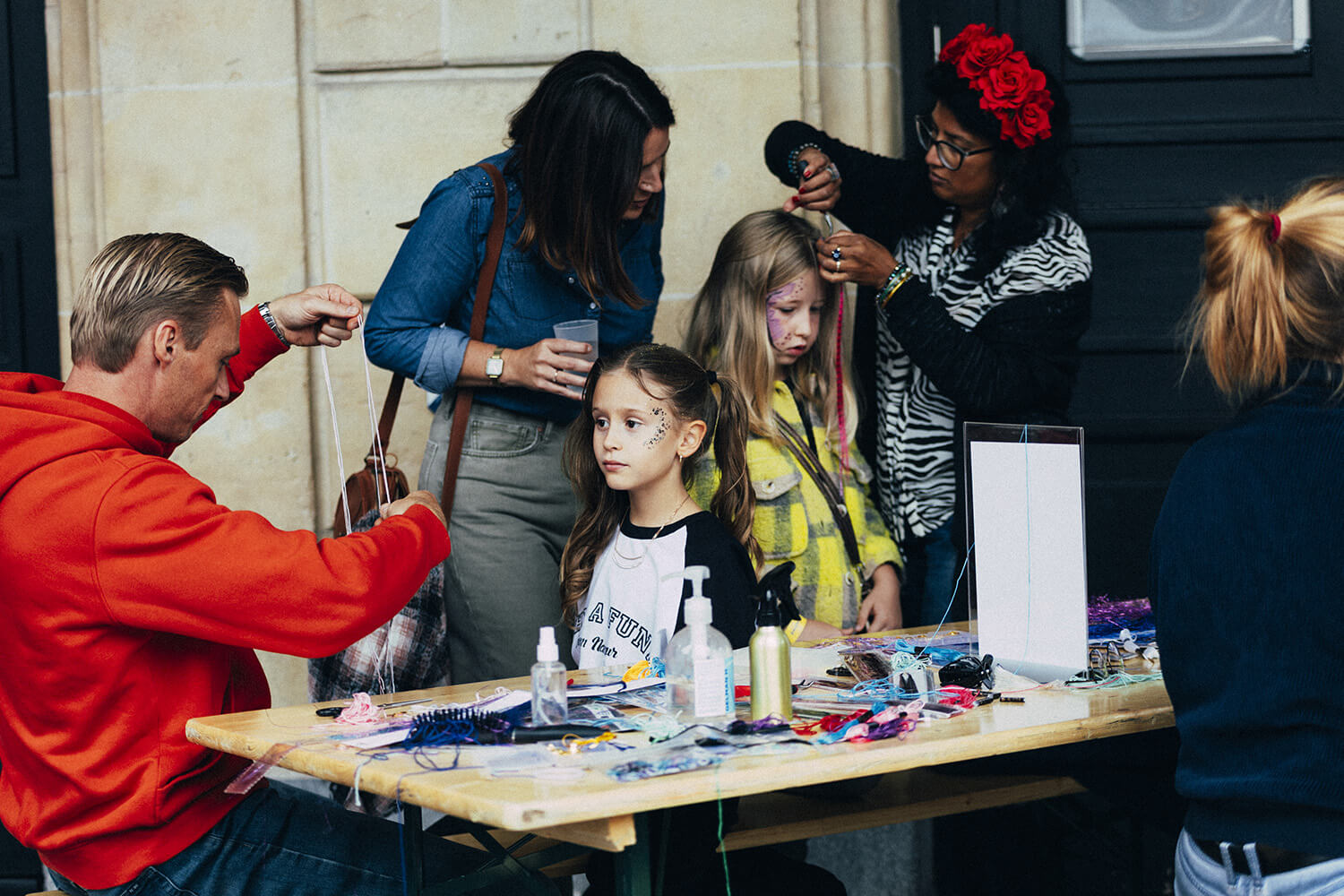 Une enfant reçoit une coiffure festive avec des accessoires colorés. Une femme, portant une couronne de fleurs rouges, arrange les cheveux de l'enfant. D'autres enfants et adultes sont présents autour d'une table remplie de produits de beauté.