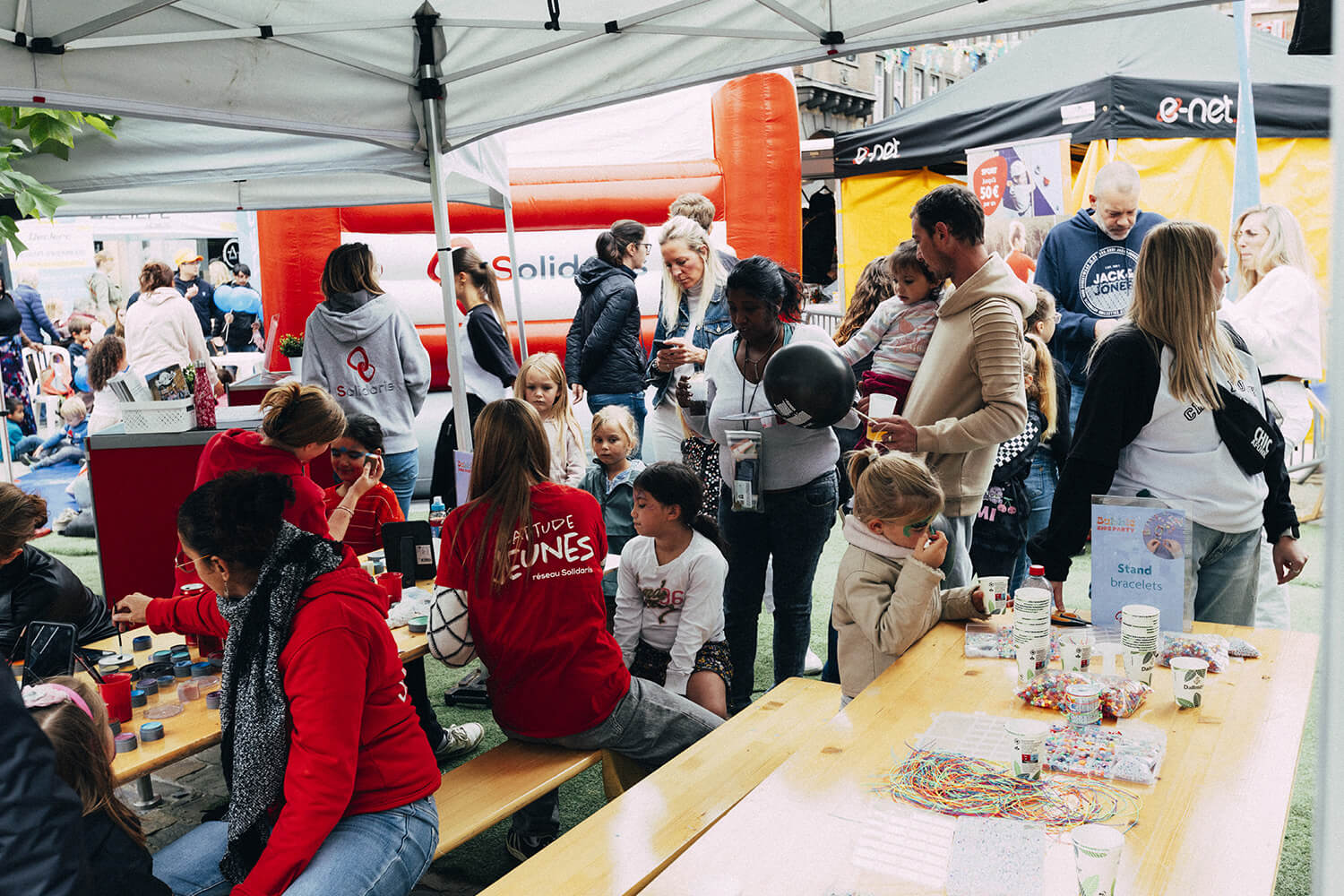 Des enfants et des adultes sont rassemblés autour d'une table d'activités lors d'un événement. Des bracelets et des accessoires sont disposés sur la table. Les enfants semblent participer à une activité manuelle ou créative.