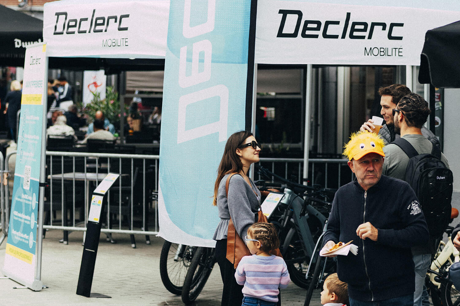 Plusieurs personnes, dont un homme portant un chapeau jaune décoré, se trouvent devant un stand marqué 