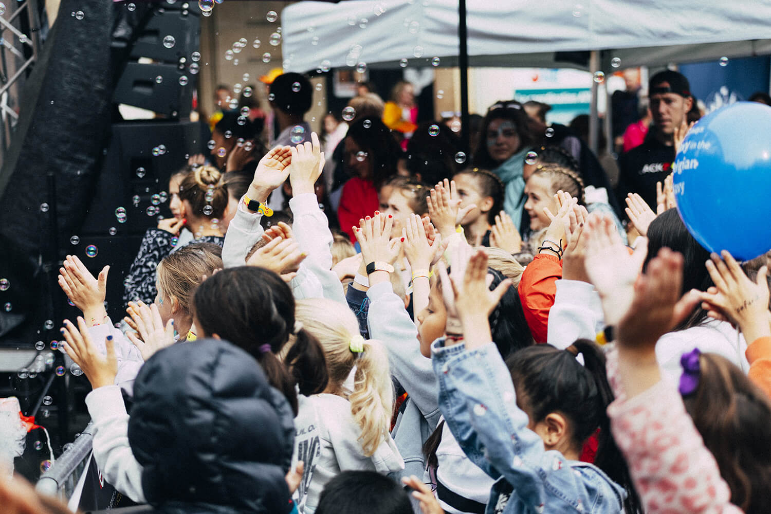 Un groupe d'enfants, les mains levées, joue avec des bulles de savon lors d'un événement en extérieur. Les bulles flottent dans l'air et un ballon bleu est visible à droite. Les enfants semblent ravis et engagés dans l'activité.