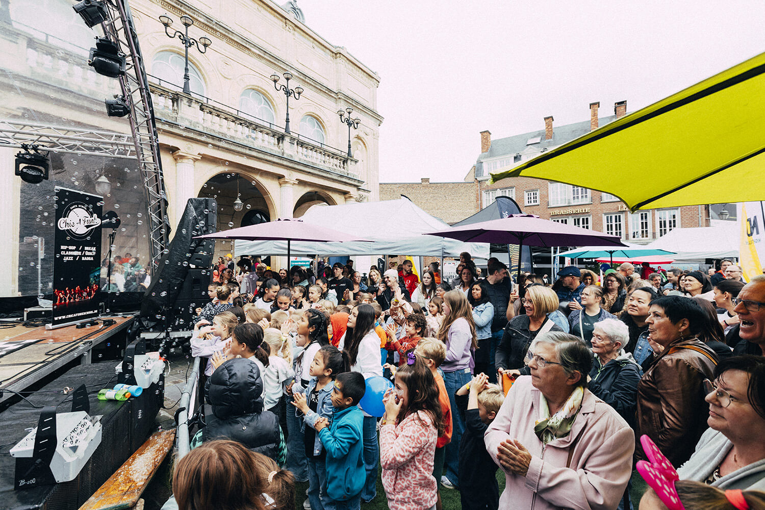 Une foule d'enfants et d'adultes se tient devant une scène extérieure lors d'un événement festif. Les enfants, au premier plan, lèvent les mains et semblent applaudir ou participer activement.