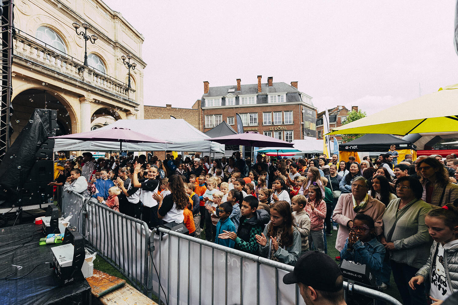 Une foule d'enfants et d'adultes se tient devant une scène extérieure lors d'un événement festif. Les enfants, au premier plan, lèvent les mains et semblent applaudir ou participer activement.