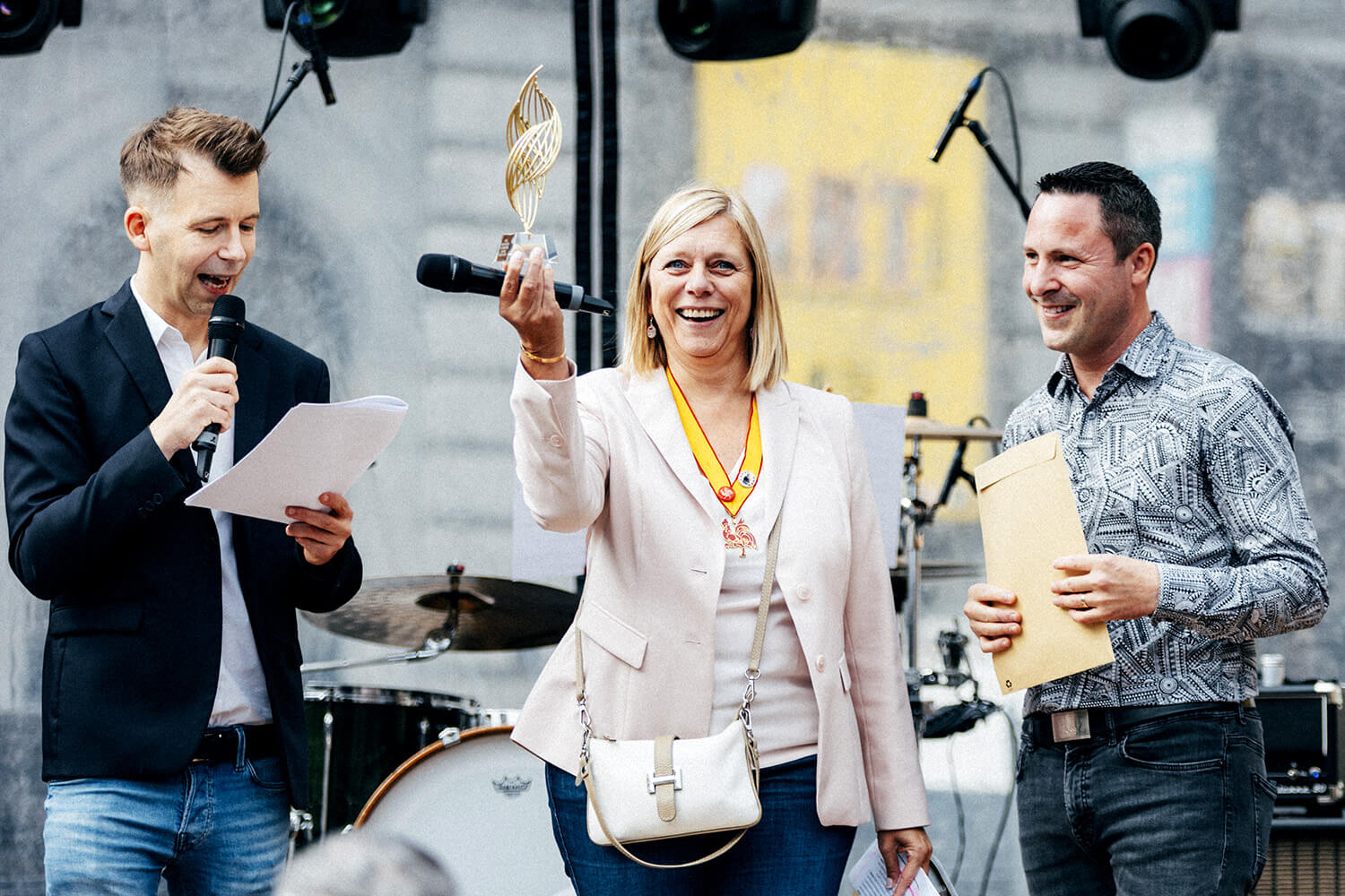 Éliane Tillieux, lauréate, souriante, lève son trophée sur scène. À ses côtés, un homme tient un micro et un document, tandis qu’un autre lauréat sourit en arrière-plan.