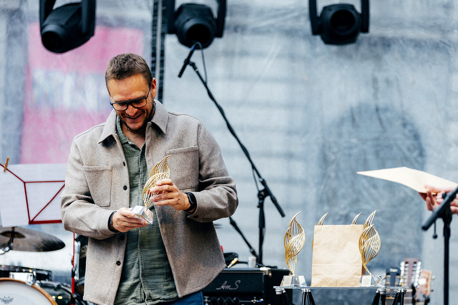 Un homme en veste grise reçoit un trophée du Prix du Souffle de l’Innovation. Il observe son prix avec émotion, entouré de matériel de scène et de projecteurs.