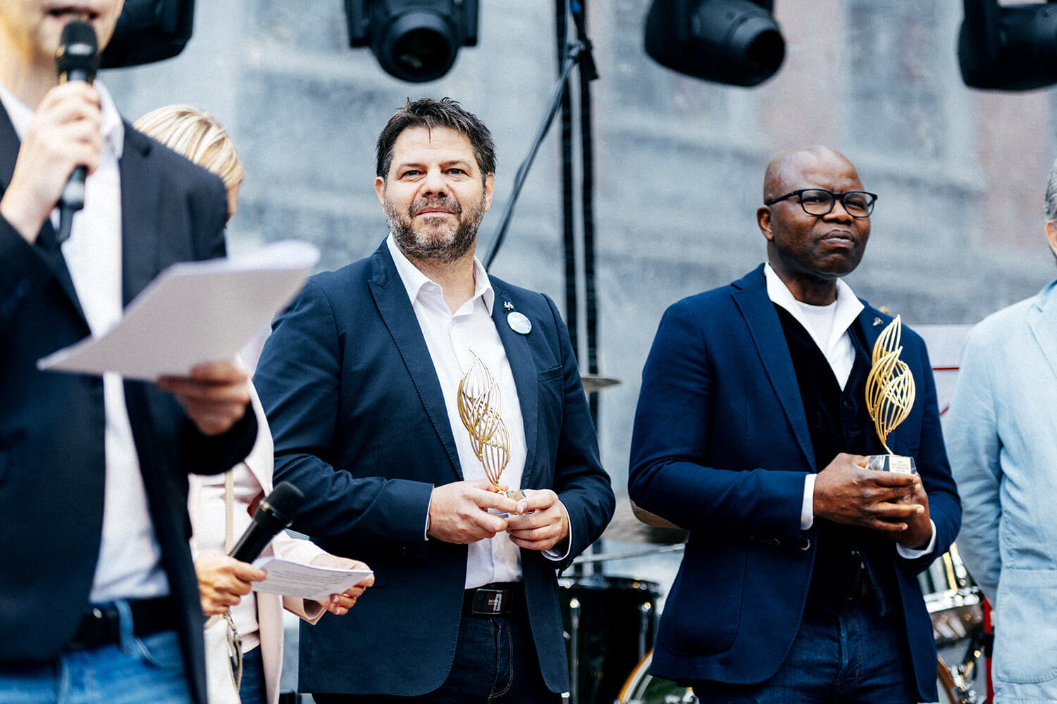 Deux hommes en costume, lauréats du Prix du Souffle de l’Innovation, tiennent chacun un trophée. Un animateur avec micro et document les accompagne sur scène.