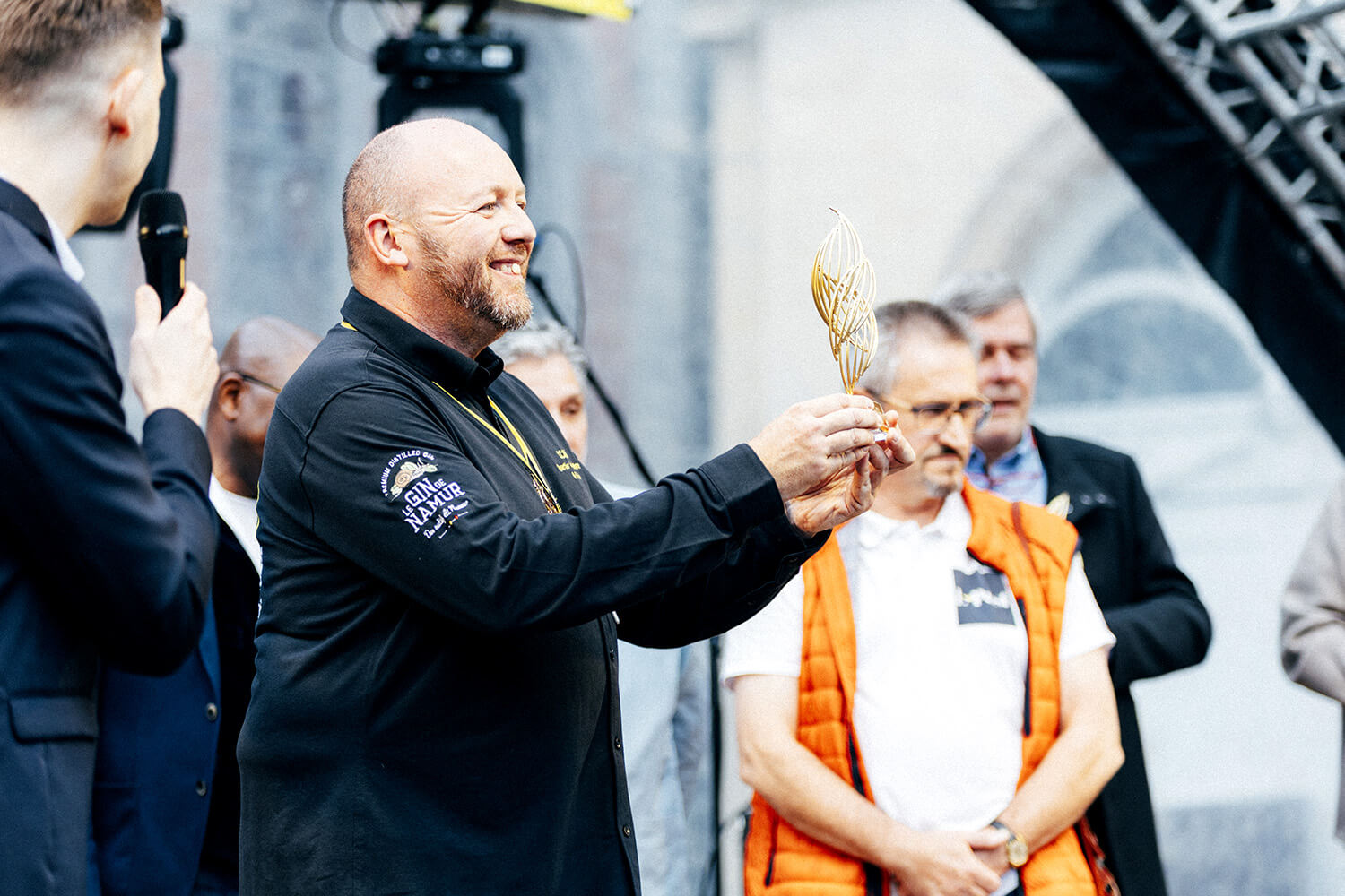 Un homme, lauréat du Prix du Souffle de l’Innovation, tient un trophée sur scène. Il sourit et regarde son prix, entouré de participants et d’un animateur avec micro.