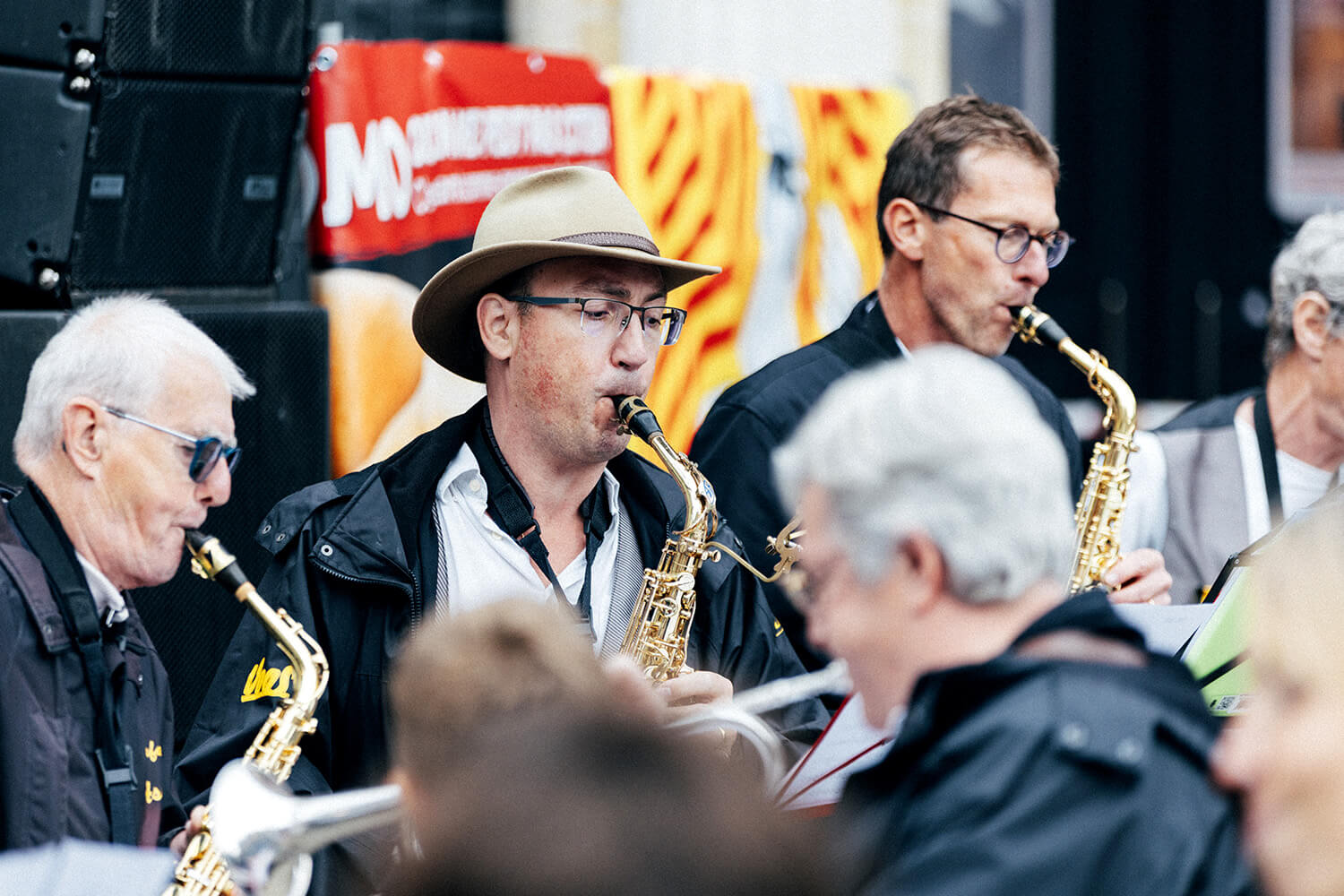 Un saxophoniste de la fanfare d’Assesse, coiffé d’un chapeau, joue avec enthousiasme lors d’une performance en extérieur. Autour de lui, d’autres musiciens et des spectateurs profitent de ce moment festif.