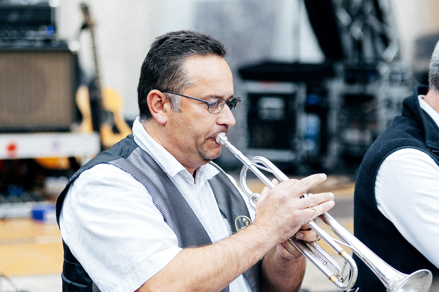 Un trompettiste de la fanfare d’Assesse interprète le morceau avec passion, vêtu d’une tenue élégante. La scène est éclairée par des projecteurs, mettant en valeur l’intensité de la performance live.