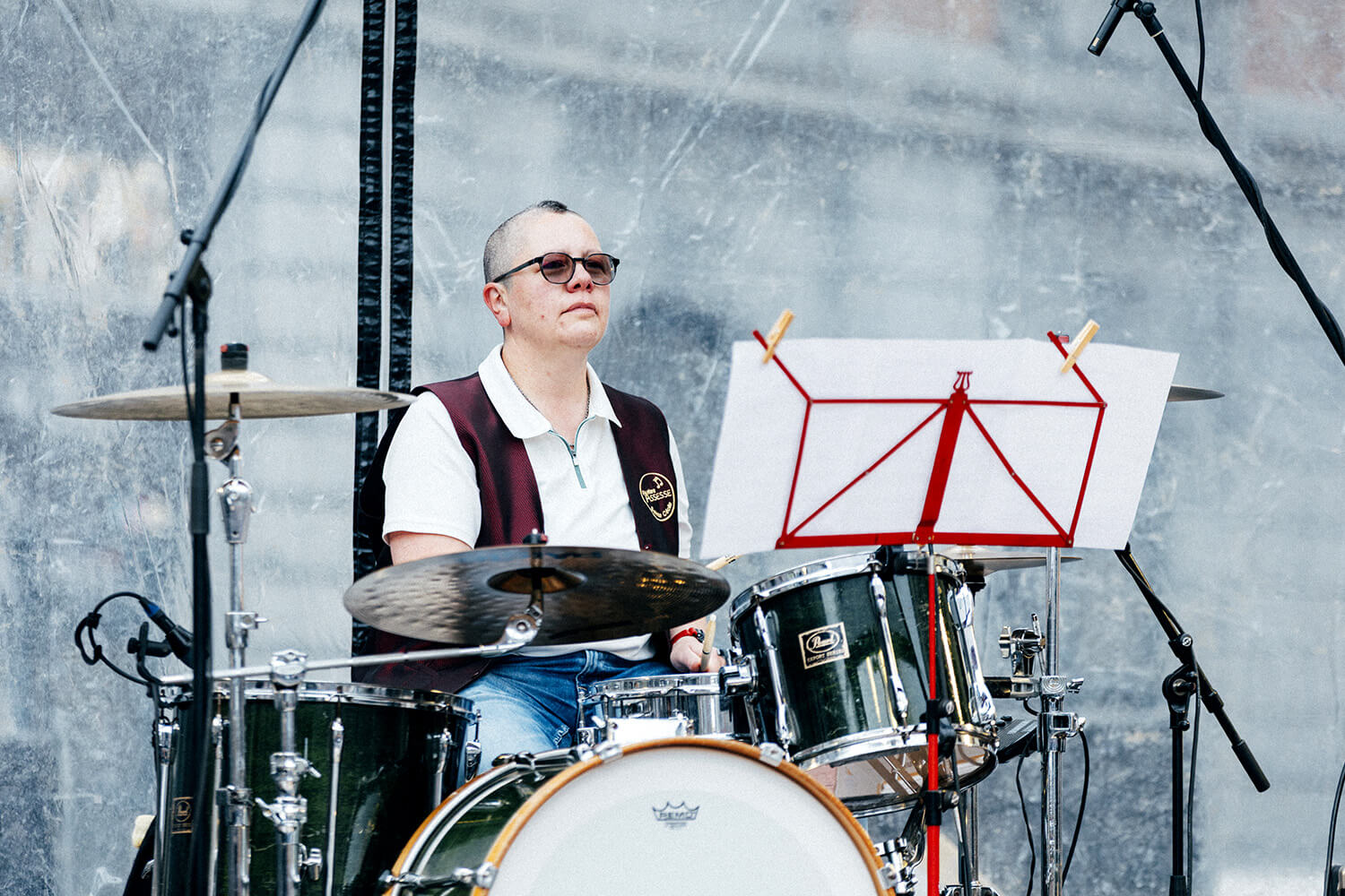 Un batteur de la fanfare d’Assesse joue avec passion sur scène, vêtu d’une chemise blanche et d’un gilet noir. Il est entouré d’équipements musicaux et de projecteurs, illustrant l’énergie live de la performance.
