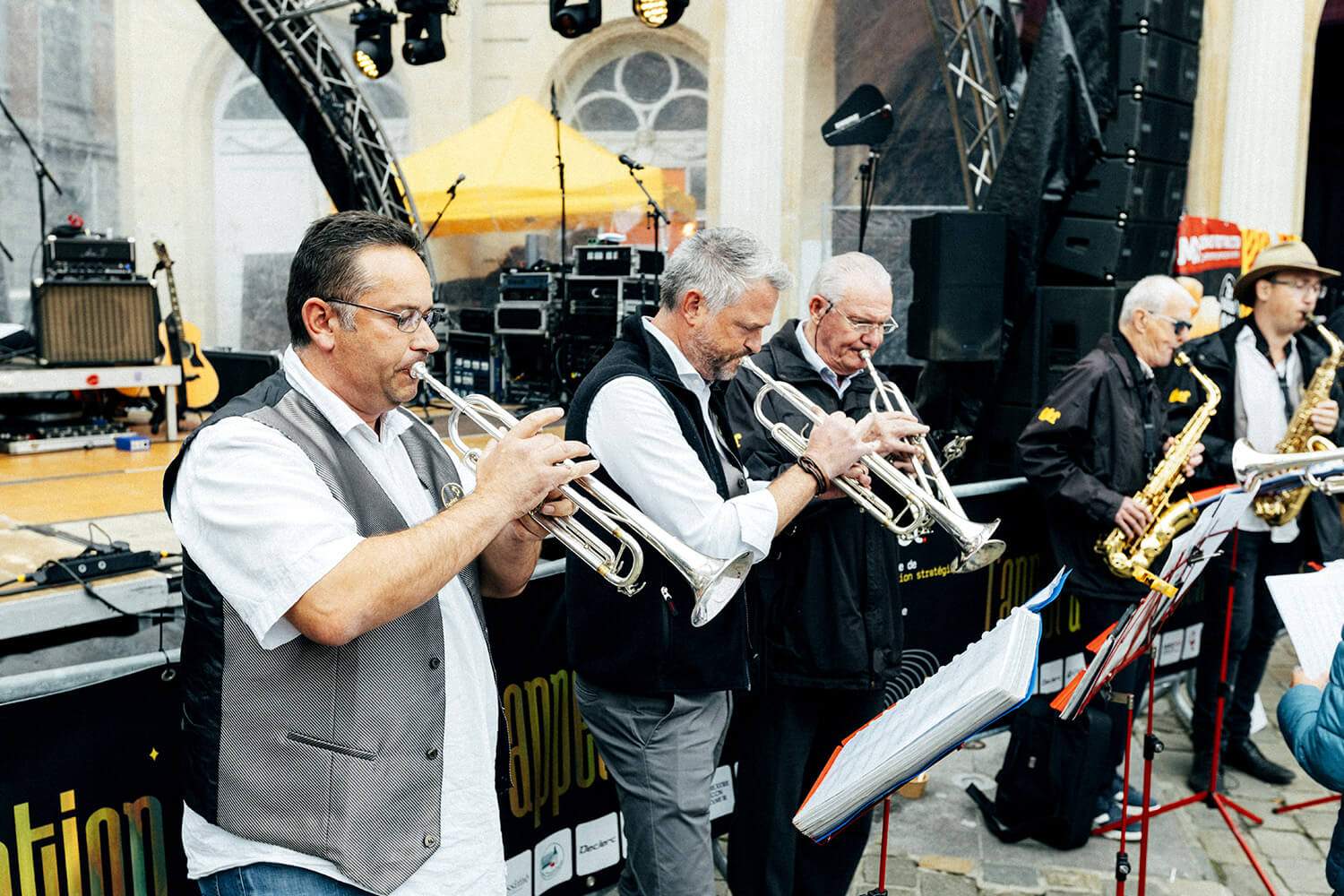 Un trompettiste de la fanfare d’Assesse joue avec fougue sur scène, entouré de ses collègues musiciens et d’équipements audio
