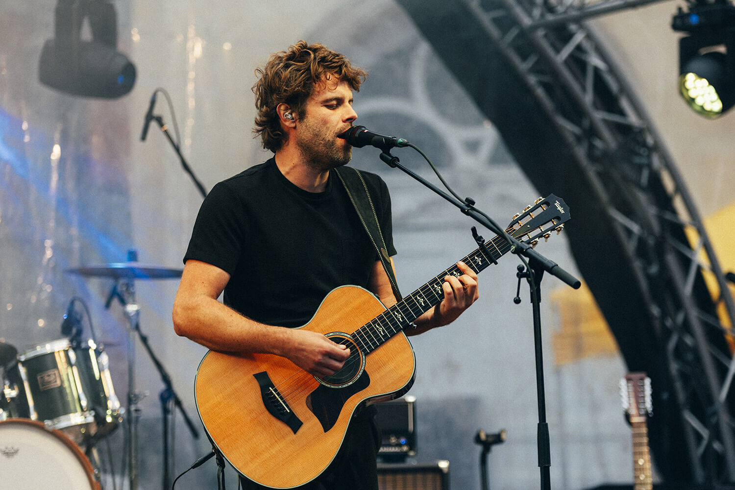 Corentin Simon, chanteur namurois, interprète une chanson en jouant de la guitare acoustique sur scène. Il porte un t-shirt noir et chante avec émotion, entouré de microphones et d’instruments de musique, dans une ambiance intimiste et lumineuse.