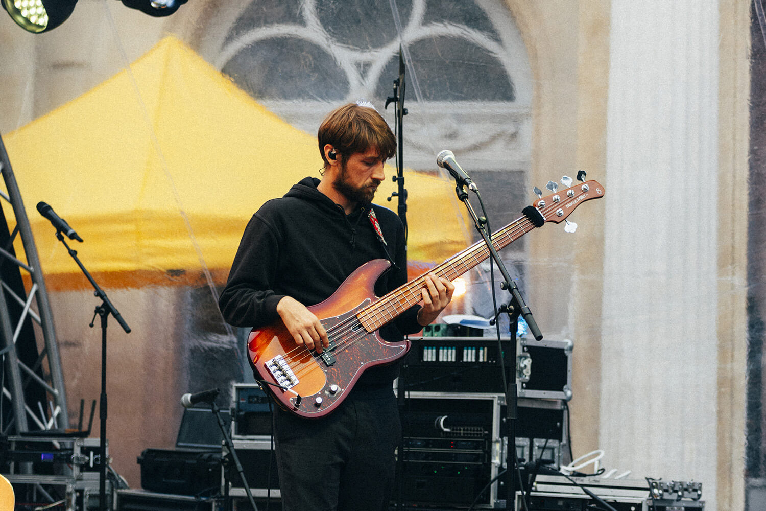 Un bassiste du trio de Corentin Simon joue de sa basse électrique sur scène, vêtu d’un sweat noir. Il est entouré de micros et d’équipements, dans une ambiance de concert en extérieur sous une tente.
