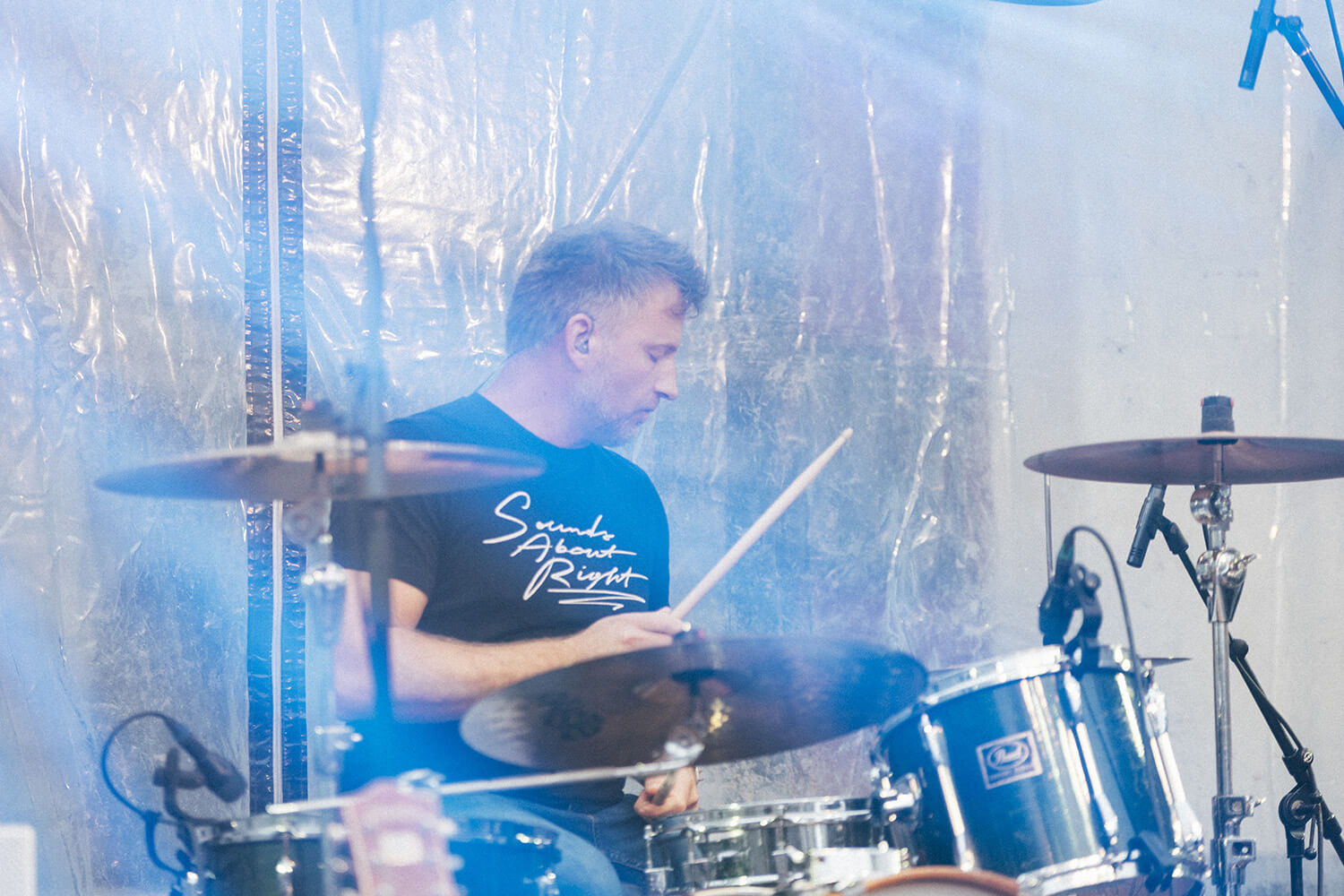 Le batteur du trio de Corentin Simon joue de la batterie sur scène, vêtu d’un t-shirt noir. Il est concentré sur sa performance, entouré de paravents transparents et de matériel de scène, dans une ambiance de concert live.