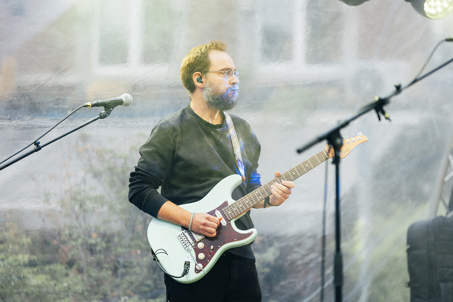 Un musicien jouant de la guitare électrique sur scène. Il porte des lunettes et un micro est positionné devant lui.