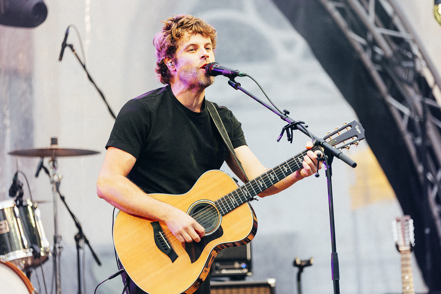 Corentin Simon chante avec passion au micro, tout en jouant de la guitare acoustique. Il est sous les projecteurs, dans une ambiance de concert intimiste et chaleureuse.
