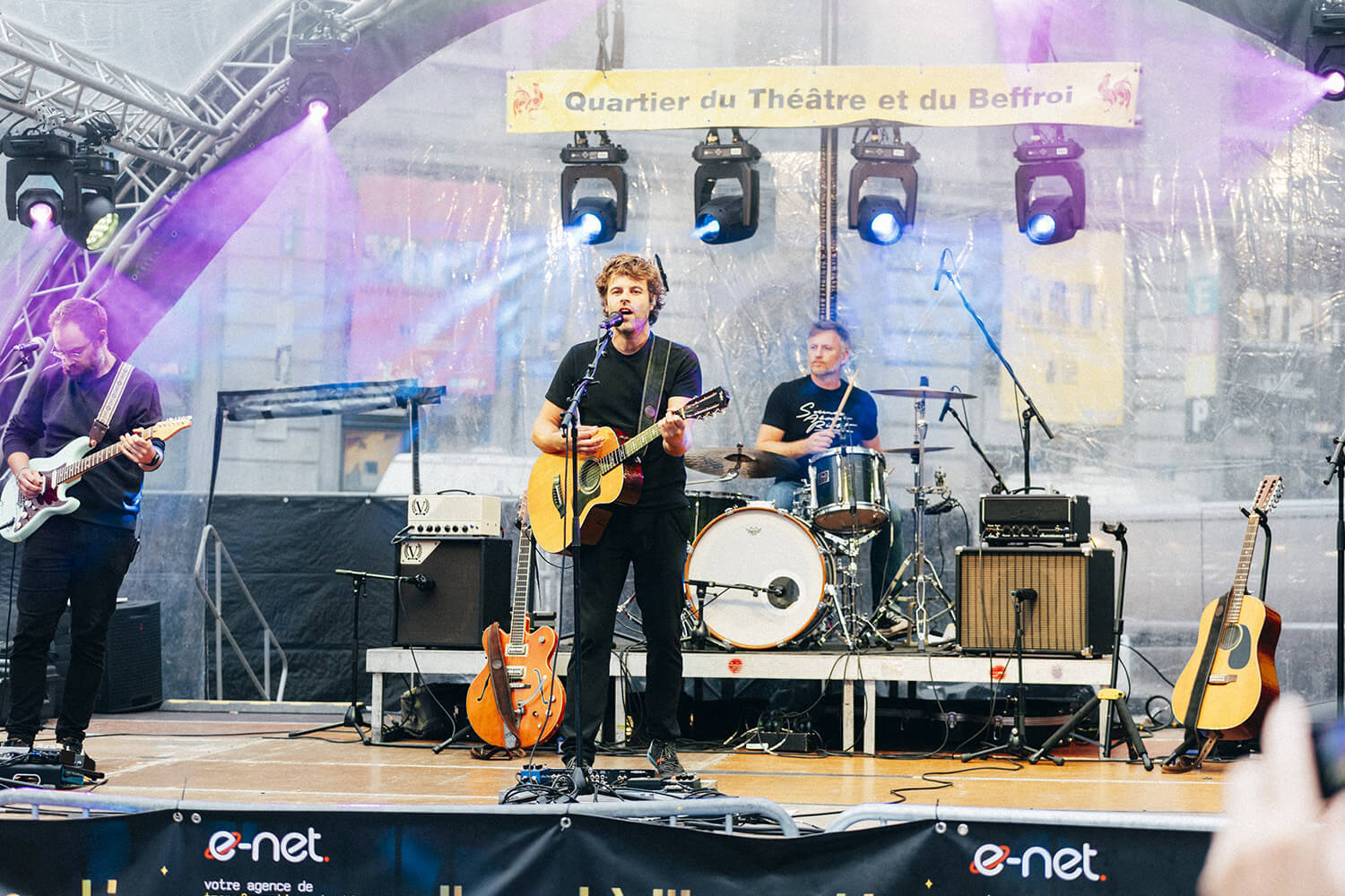 Corentin Simon et son trio live jouent sur scène lors d'un concert en extérieur. Corentin chante et joue de la guitare acoustique, entouré d'un guitariste électrique et d'un batteur.