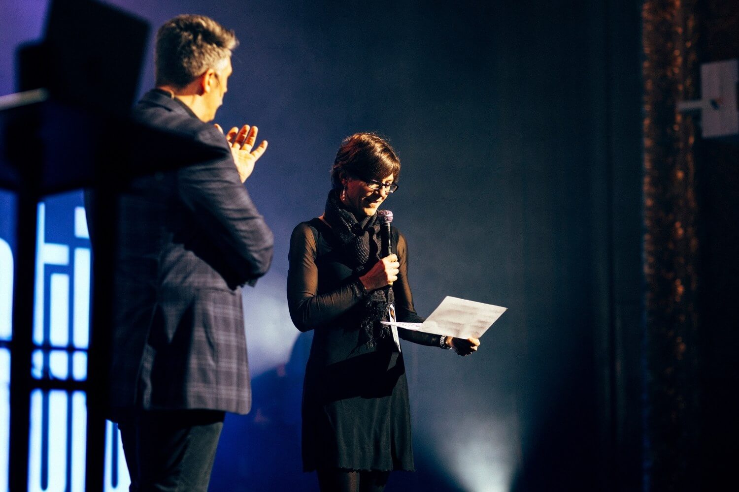 Une participante lit un texte sur scène, accompagnée par Benjamin Gishlain.
